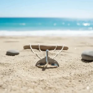 Coastal lifestyle image of the whale tail necklace lying on beach sand with the ocean in the background, reinforcing its nautical theme.