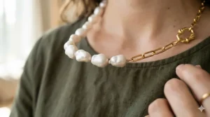 A detailed macro close-up shot focusing on the transition point of an asymmetrical necklace, showing organic textured baroque pearls meeting a sleek gold paperclip chain link on a green linen shirt.